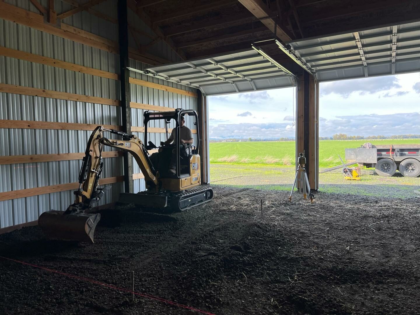 Excavator grading and leveling gravel inside a barn for shed pad preparation and sitework.